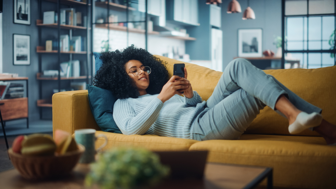 Happy Beautiful Latina Female Using Smartphone in Cozy Living Room at Home. Female Resting on Comfortable Sofa. She’s Browsing the Internet and Checking Videos on Social Networks and Having Fun.